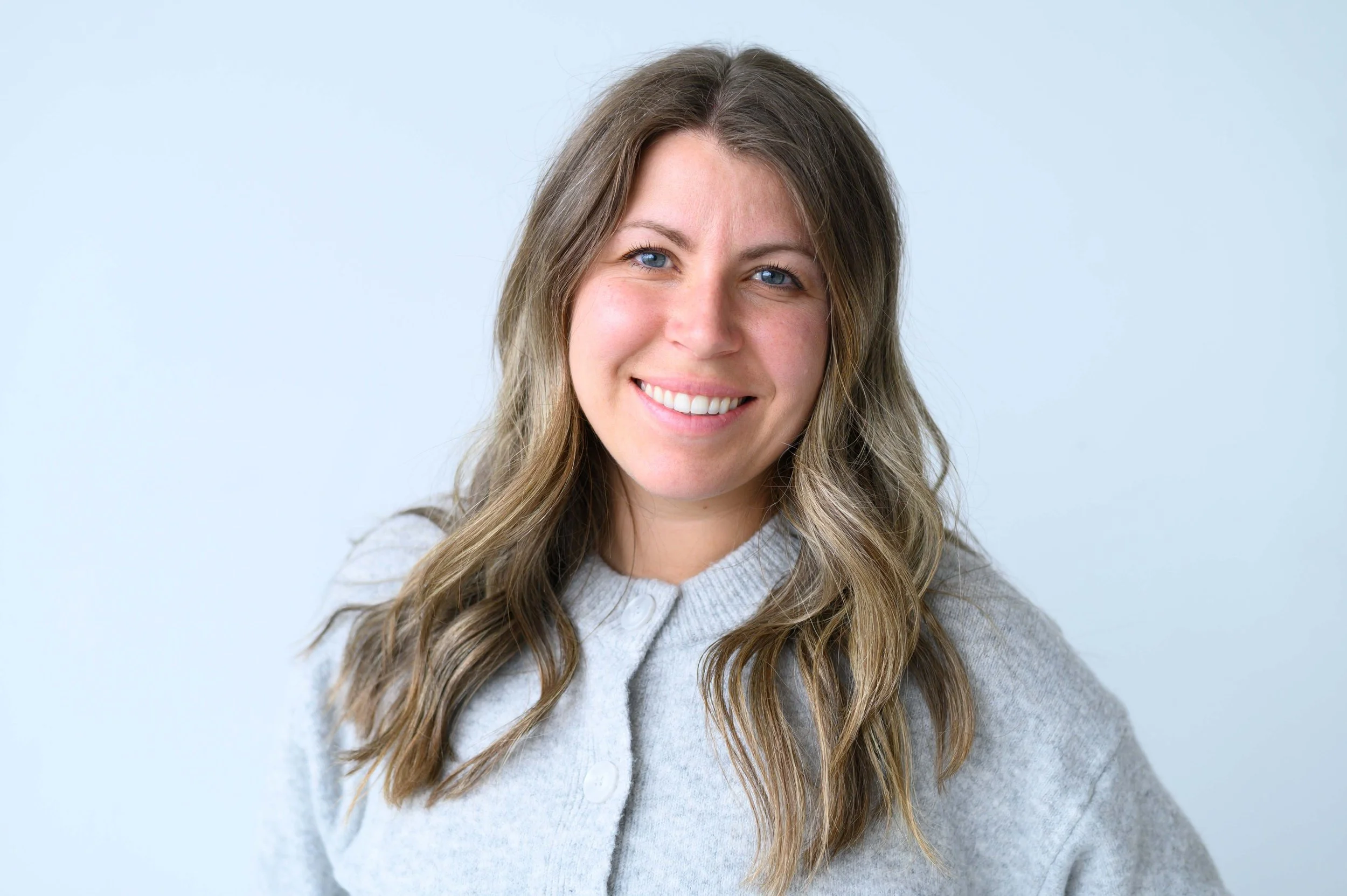 Jeune femme souriante avec des cheveux longs et ondulés, portant un pull gris clair, contre un fond blanc clair.