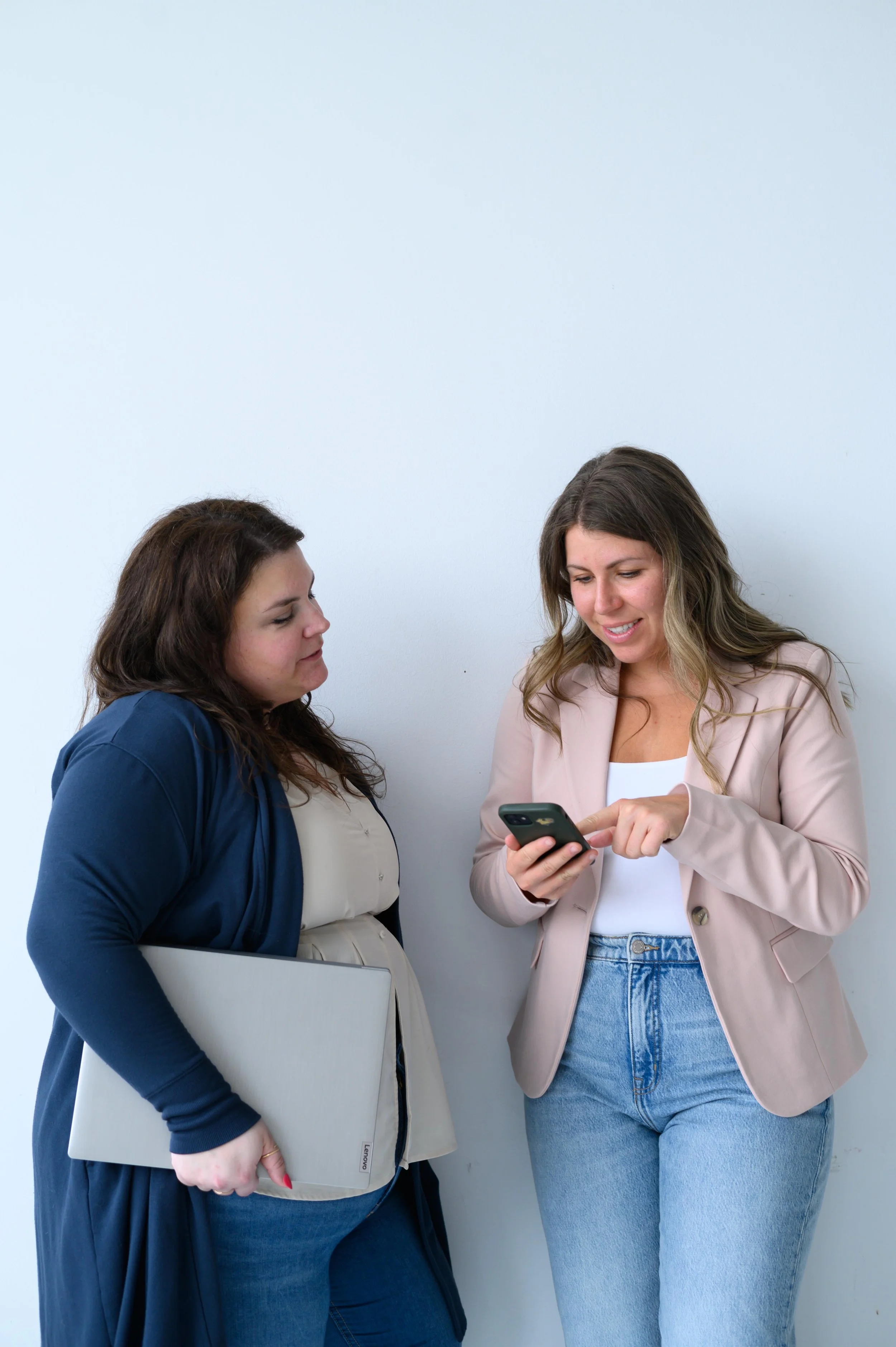 Deux femmes discutent et regardent un téléphone portable, l'une tenant un ordinateur portable. Le fond est blanc.