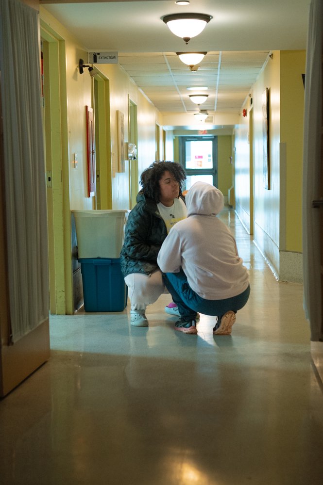 Deux personnes dans un couloir d'immeuble, l'une agenouillée, située face à l'autre qui est également agenouillée, apparemment lors d'une conversation ou d'une assistance.