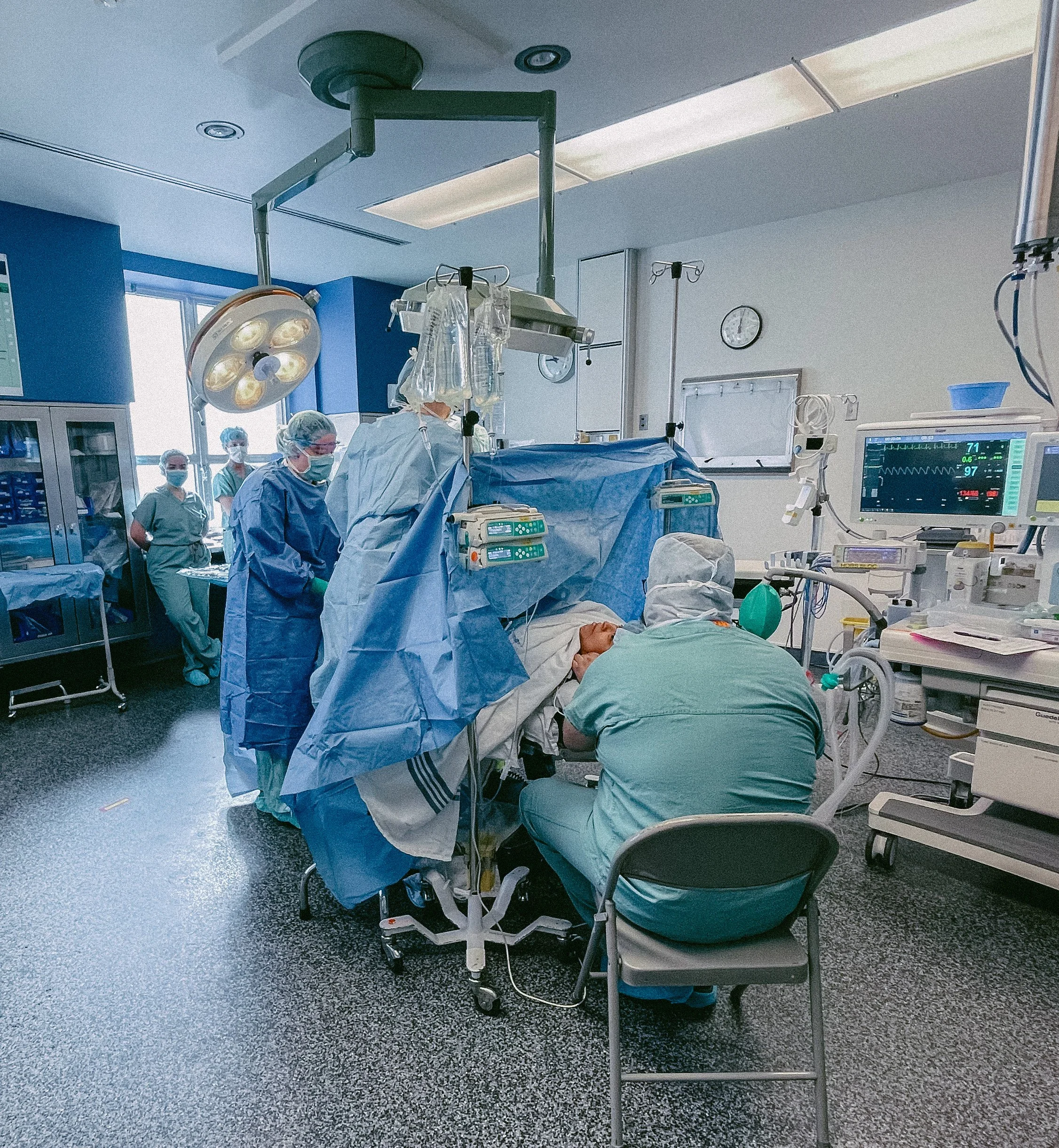 Salle d'opération chirurgicale avec médecins en tenue stérile, patient sur la table sous des draps chirurgicaux, équipement médical et moniteurs.