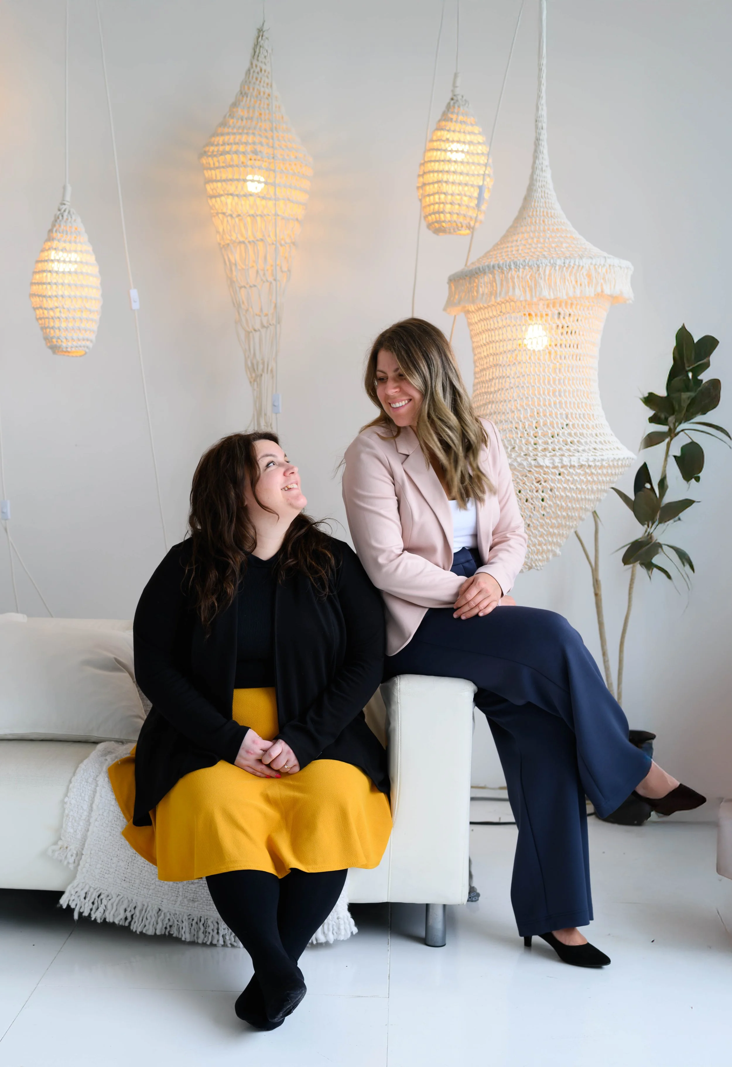 Deux femmes souriantes assises sur un canapé blanc, avec des lampes en macramé suspendues derrière elles, dans une pièce lumineuse et décorée avec un plante verte.