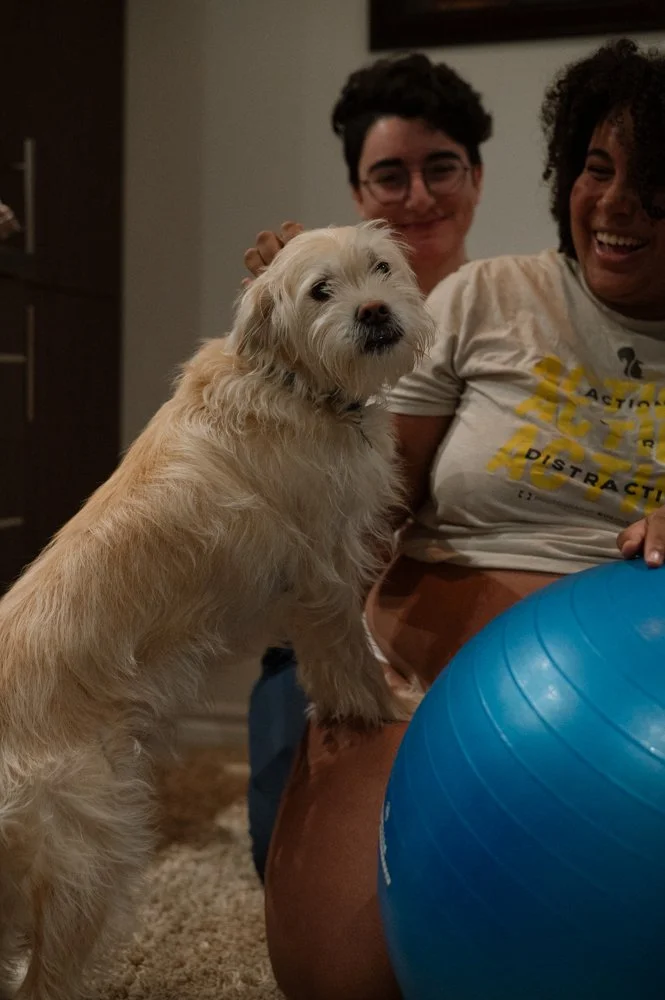 Deux femmes souriantes avec un chien beige à la patte sur leur genou, tenant un ballon bleu dans une pièce intérieure.