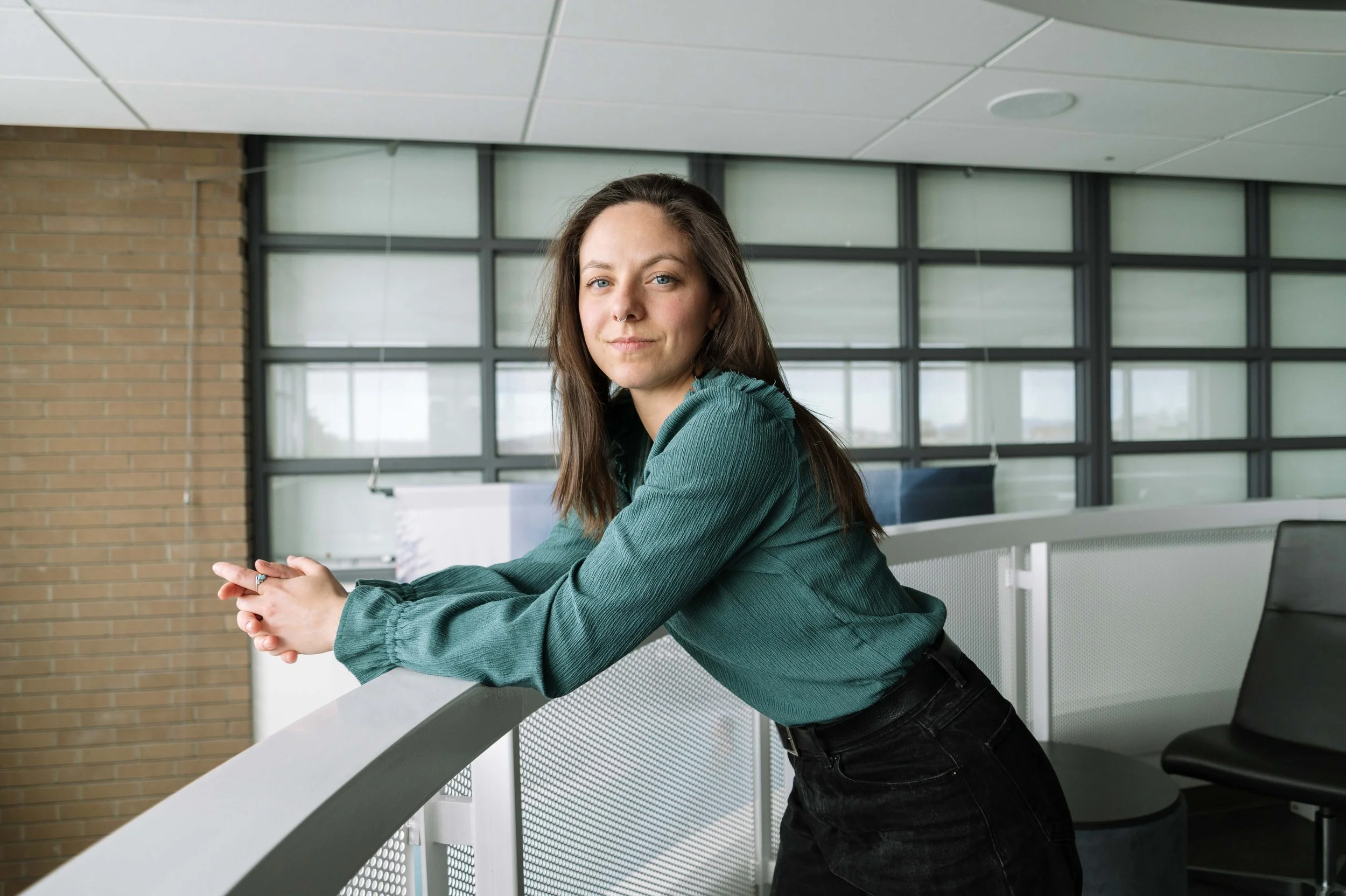 Jeune femme décontractée, posant dans un espace moderne, portant une chemise verte et un pantalon noir, dans un bâtiment lumineux avec des fenêtres et une décoration minimaliste.