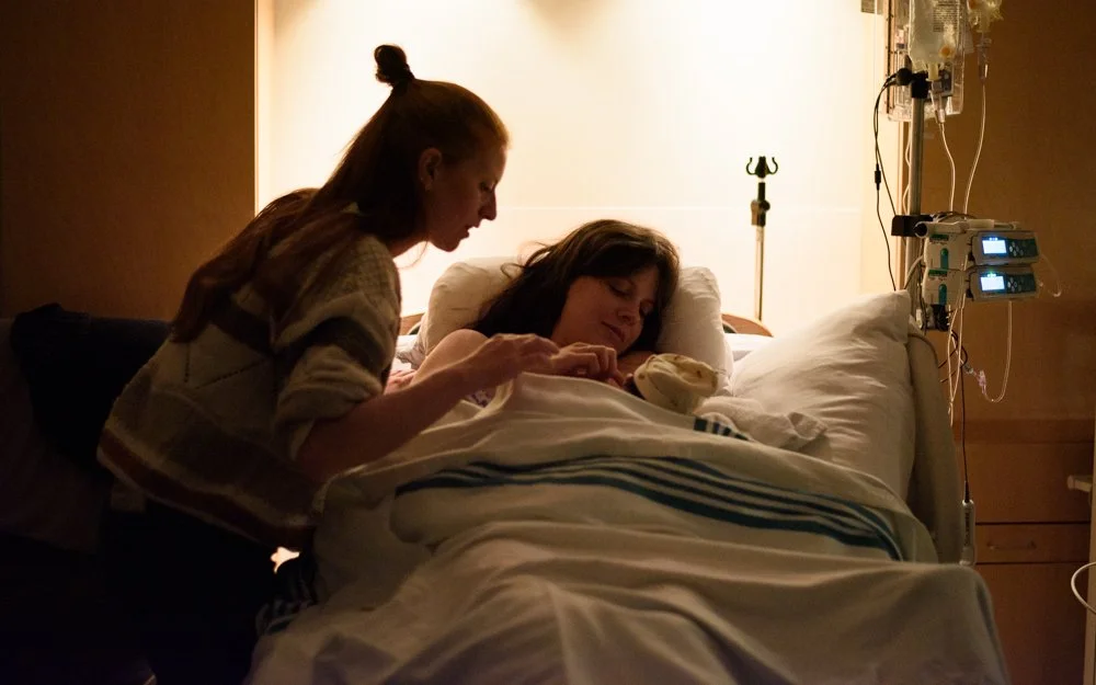 Une femme dans un lit d'hôpital, apparemment malade ou en convalescence, avec une autre femme près d'elle, peut-être une proche ou une infirmière, dans une pièce éclairée doucement semble-t-il. La scène évoque un moment de soins ou de soutien.