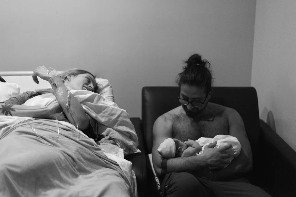 Une femme à l'hôpital, allongée sur un lit, sourit en recevant une perfusion. Un homme sans chemise, portant des lunettes, tient un nouveau-né dans ses bras, assis sur un canapé à côté du lit.