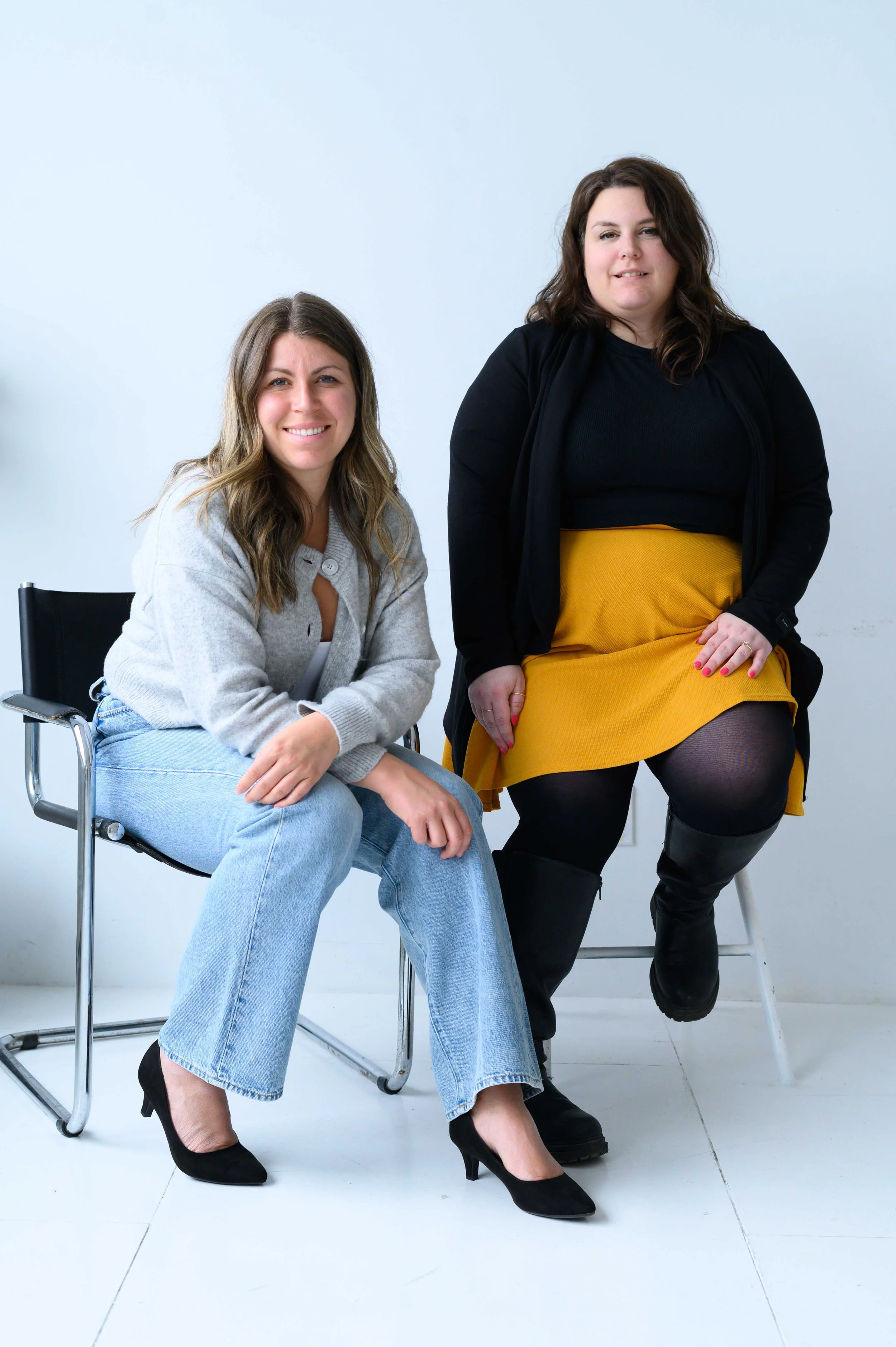 Deux femmes posent pour une photo dans une pièce blanche. L'une est assise avec un pull gris, jean et chaussures à talons noirs, l'autre est assise sur une chaise, portant une robe verte, un cardigan noir et des bottes noires.