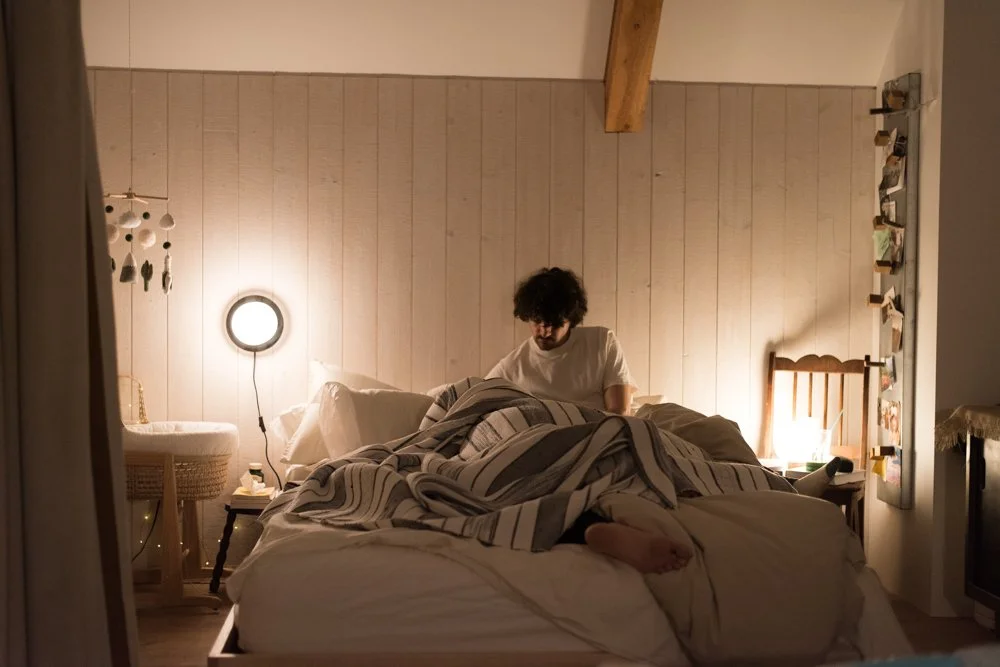 Une personne assise sur un lit dans une chambre, entourée de lampes et de décorations chaleureuses, avec un mur en bois derrière eux.