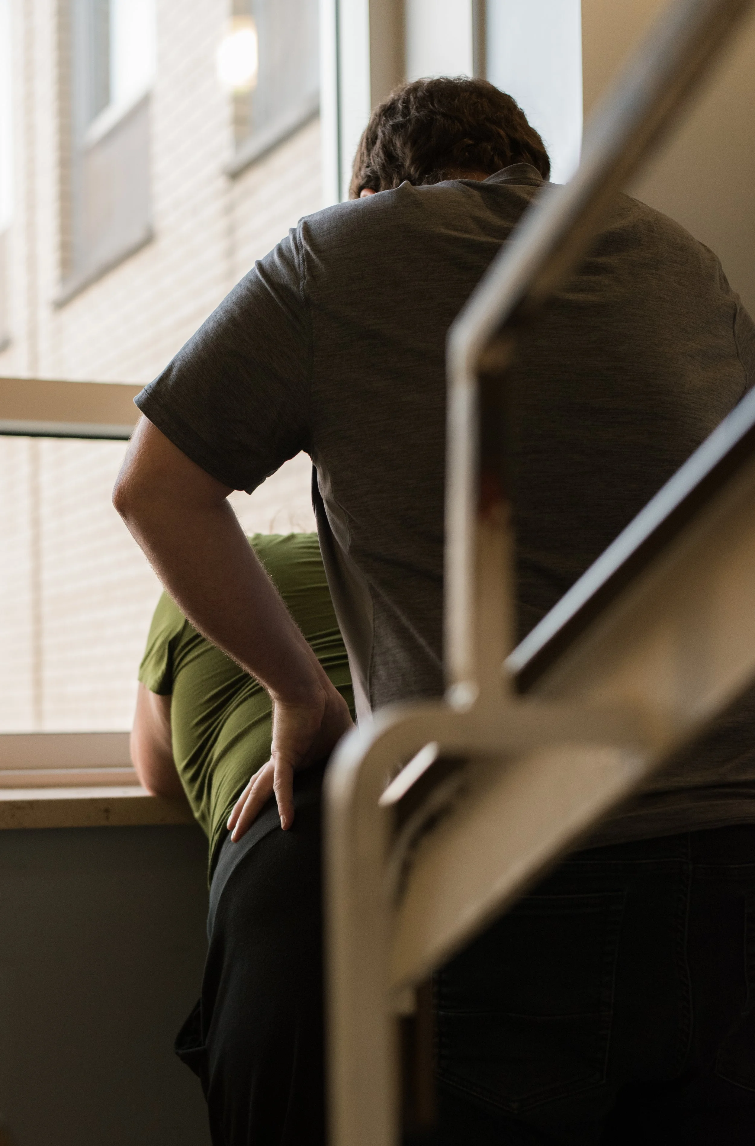 Deux personnes debout près d'une fenêtre, l'une portant un t-shirt vert et l'autre un t-shirt gris, regardant à l'extérieur.