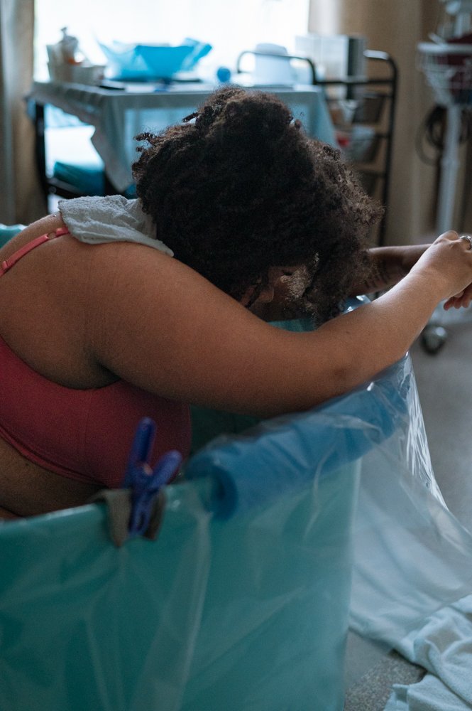 Une femme semble épuisée ou triste, assise dans une grande boîte en plastique dans une pièce d'hôpital ou clinique, avec des équipements médicaux et un lit en arrière-plan.
