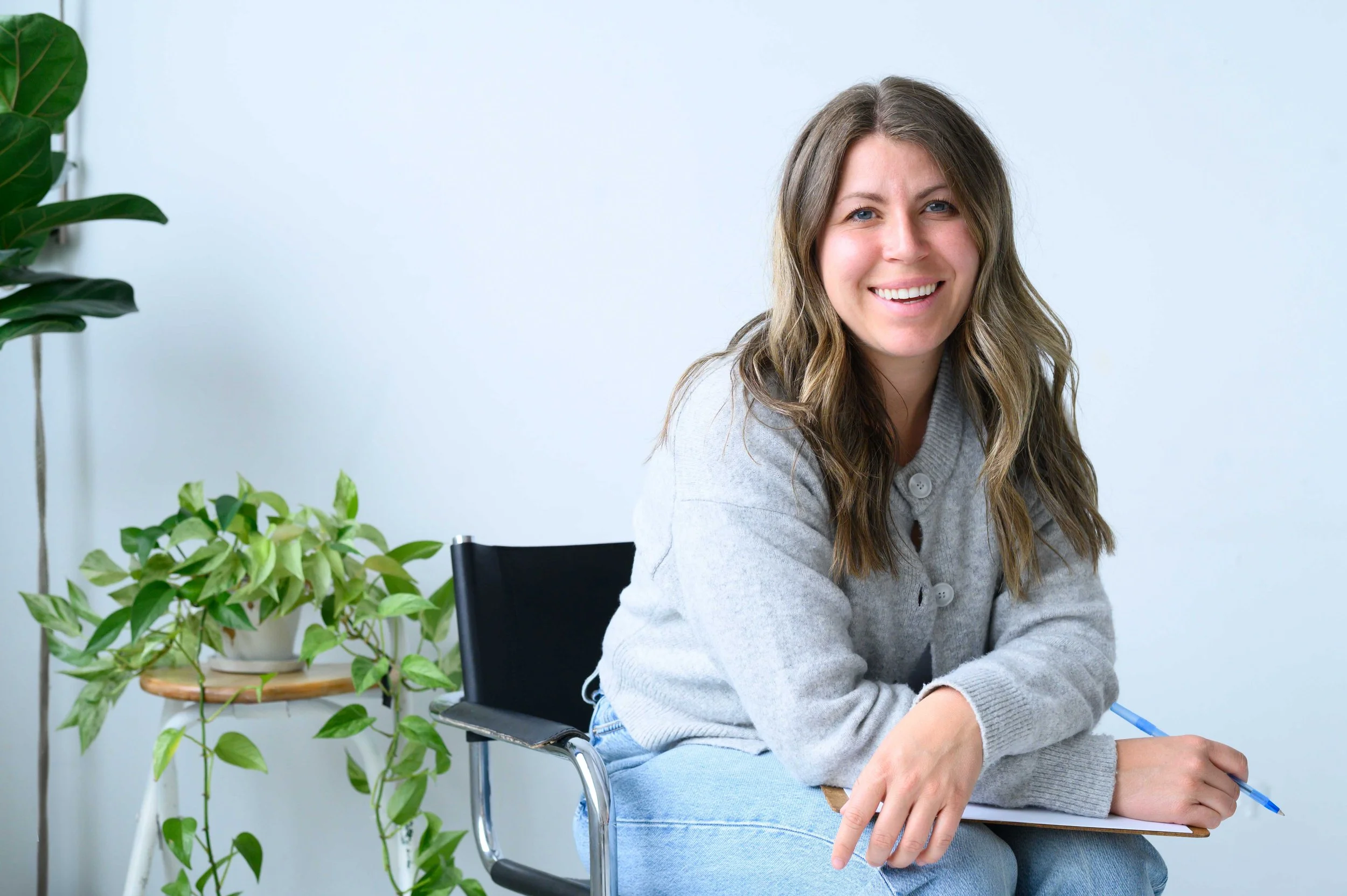 Jeune femme souriante assise en fauteuil, tenant un carnet et un stylo, avec des plantes vertes à côté, dans un intérieur lumineux.