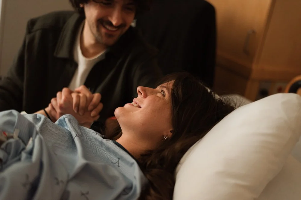 Une femme allongée dans un lit d'hôpital, souriant en regardant un homme debout à côté d'elle, qui tient ses mains. La femme semble être une patiente, peut-être récemment arrivée à l'hôpital ou après un accouchement. L'homme est probablement un proch
