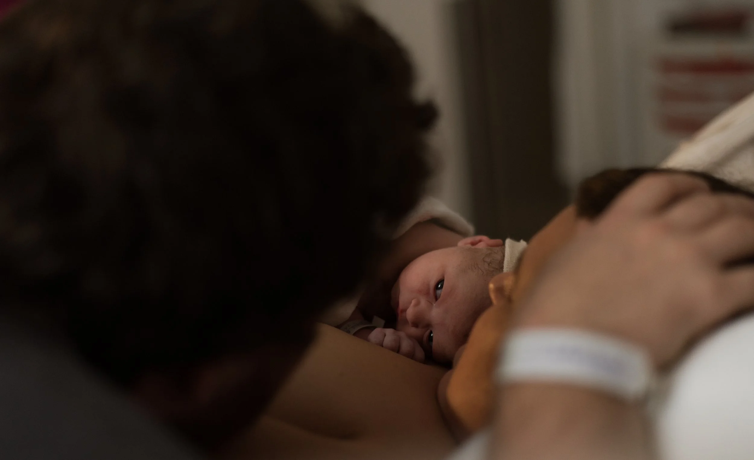 Un nouveau-né allongé sur le torse d'un adulte, couvert d'une serviette, avec une personne en arrière-plan tenant le bébé.