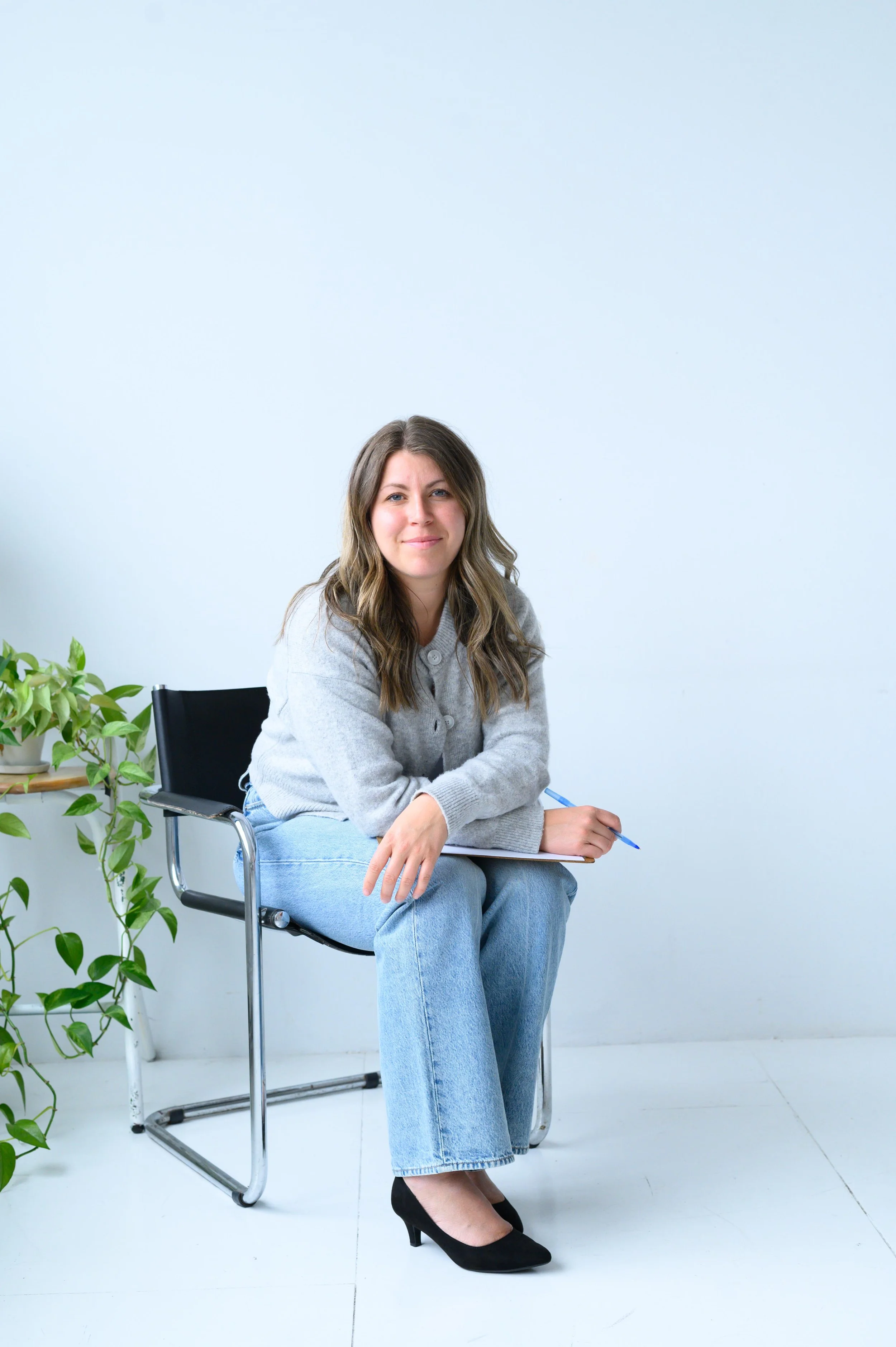Une femme assise sur une chaise noire avec un carnet et un stylo, portant un pull gris, un jean bleu et des talons noirs, dans une pièce avec un mur blanc et une plante verte à gauche.