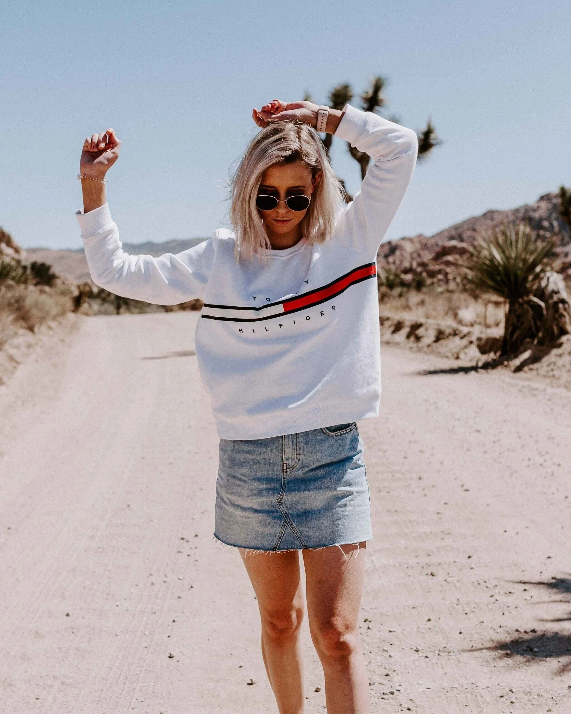 Person wearing sunglasses, white Tommy Hilfiger sweatshirt, and denim skirt on desert road