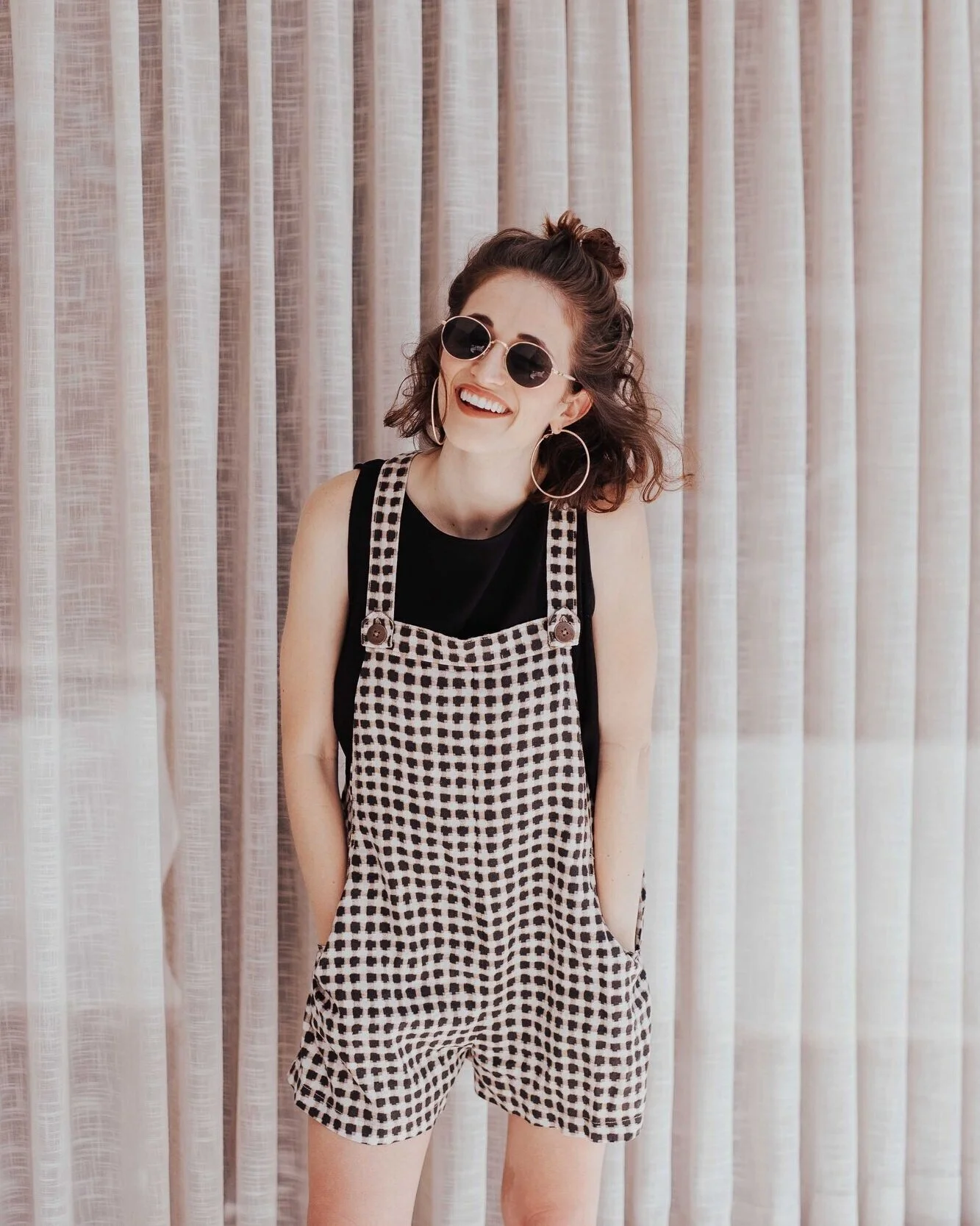 Person wearing sunglasses, large hoop earrings, black top, and checkered overalls standing in front of a curtain, smiling.