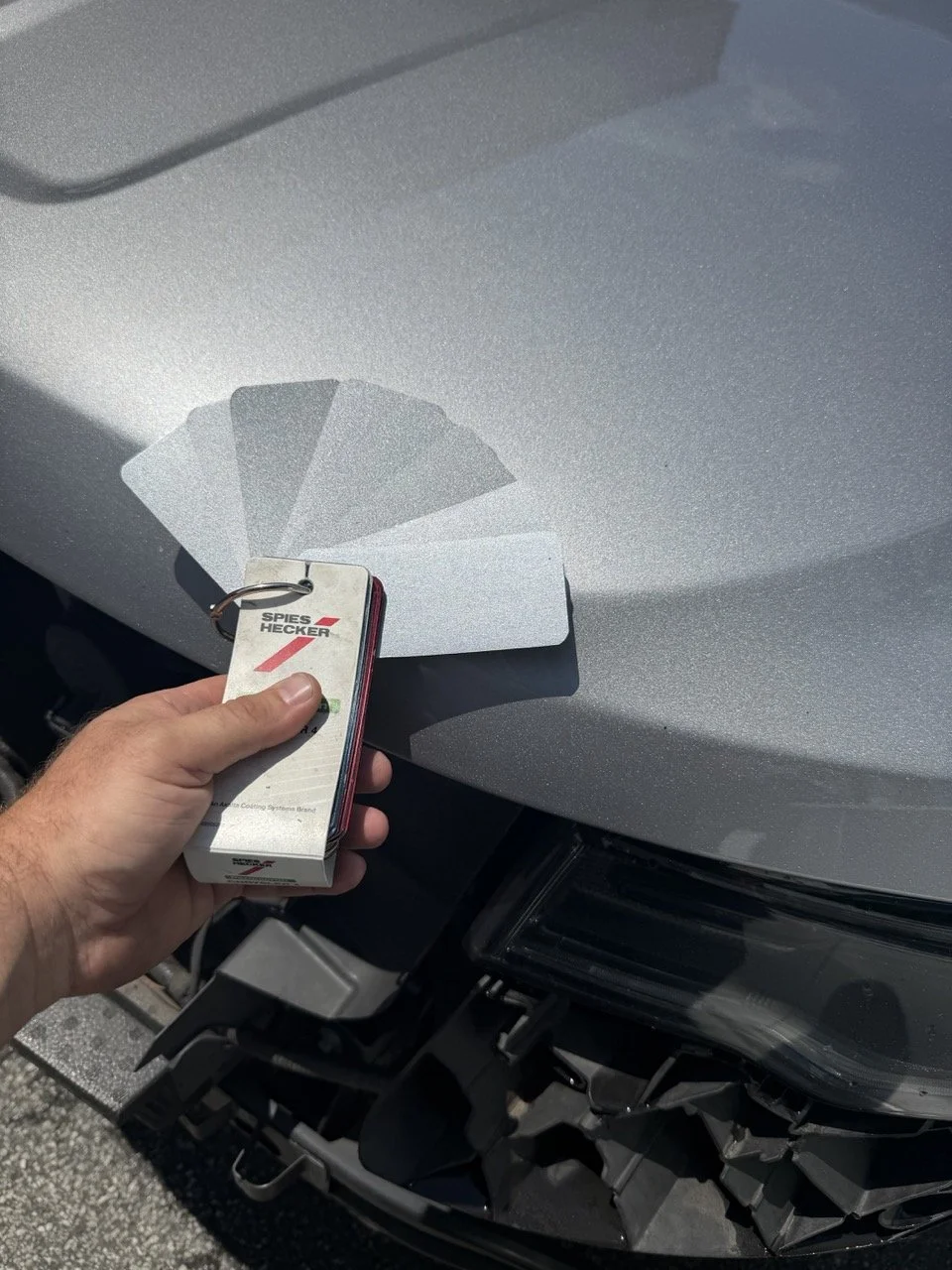 Hand holding a set of paint color samples and a keychain in front of a car's hood, likely selecting or matching paint for the vehicle.