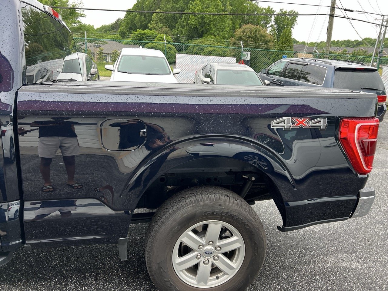 Black pickup truck with 4x4 emblem on rear side, parked in a lot with several other cars, water droplets on surface, reflected image of person taking photo, trees and a fence in background.