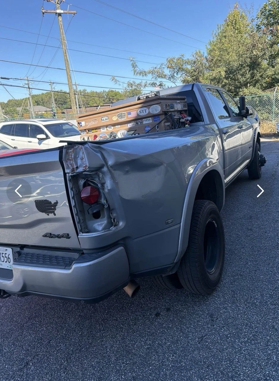 Damaged silver pickup truck with a crushed rear left side, parked in a lot with other vehicles and utility poles under a clear blue sky.