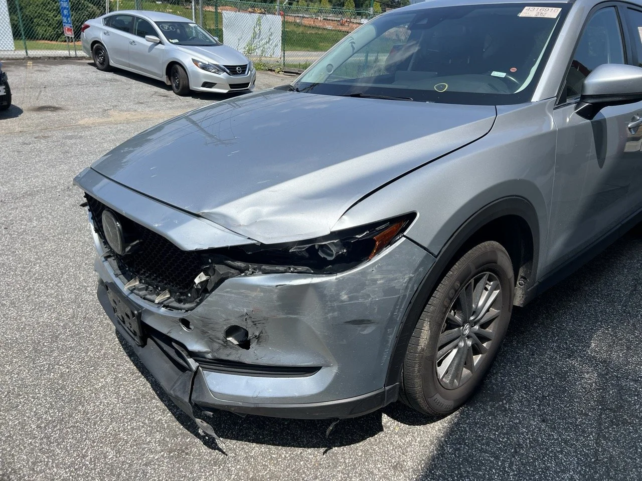 Gray car with front-end damage, missing grille, and bent hood, parked on a lot with other cars in background.