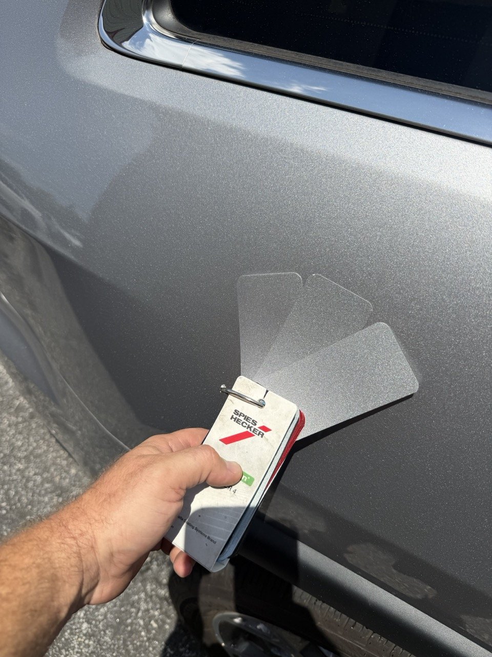 A person applying a paint protection film to the front fender of a silver car using a toolkit labeled 'SPEES HECKER' with transparent film sheets.