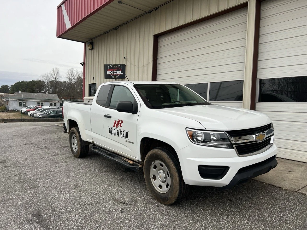 White Chevrolet pickup truck parked outside a building with closed garage doors, featuring company decals and an Excel sign above.