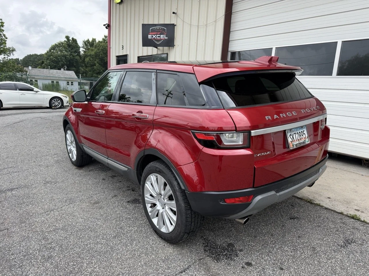 Red Range Rover SUV parked outside a building with a white garage door and an 'Excel' sign, with a white sedan in the background.