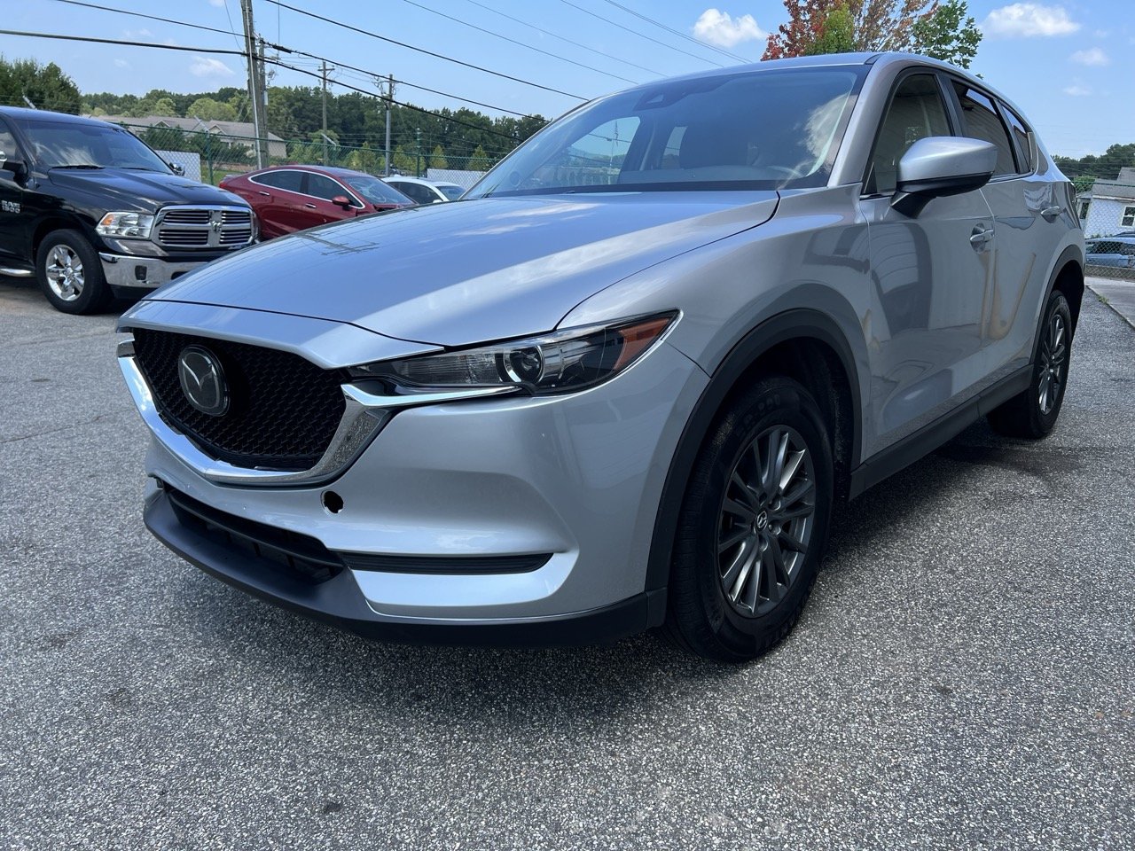 Silver Mazda CX-30 parked in a lot with other vehicles, including a black pickup truck and a red sedan, with a chain-link fence and trees in the background.