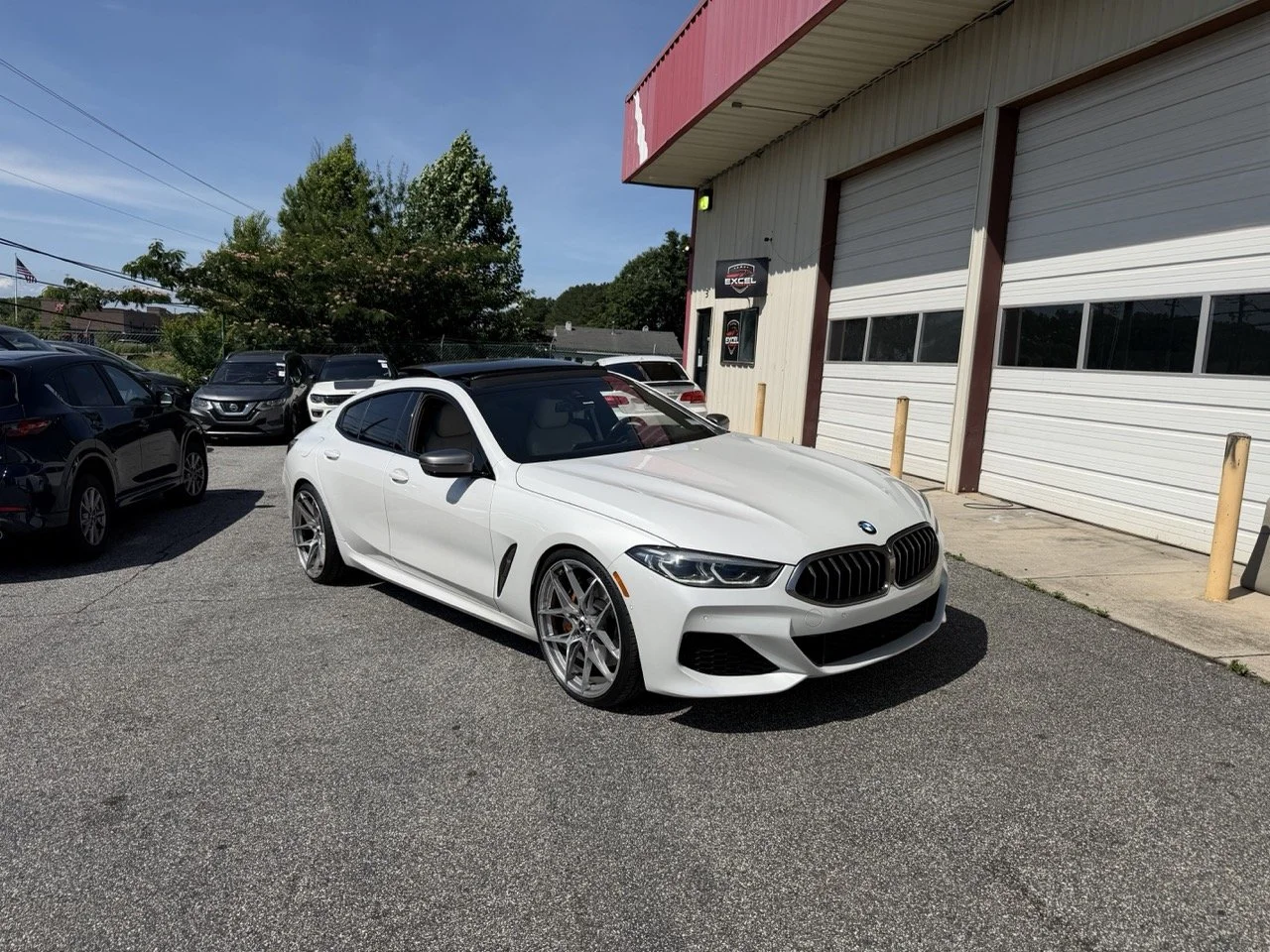A white BMW parked in a lot outside a building with a garage door, with other cars and greenery in the background.