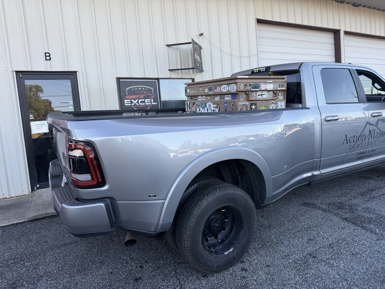 Silver pickup truck parked outside a building with signage, hauling a stack of wooden planks decorated with numerous stickers in the truck bed. The building has two roller doors and a glass door with a sign.