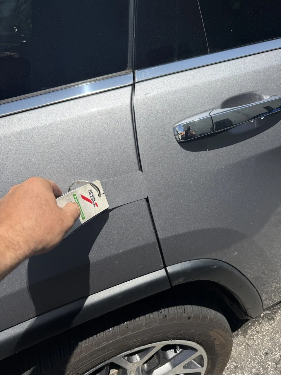 Person applying a parking permit sticker to the front left fender of a metallic gray SUV, with part of the tire and door handle visible.