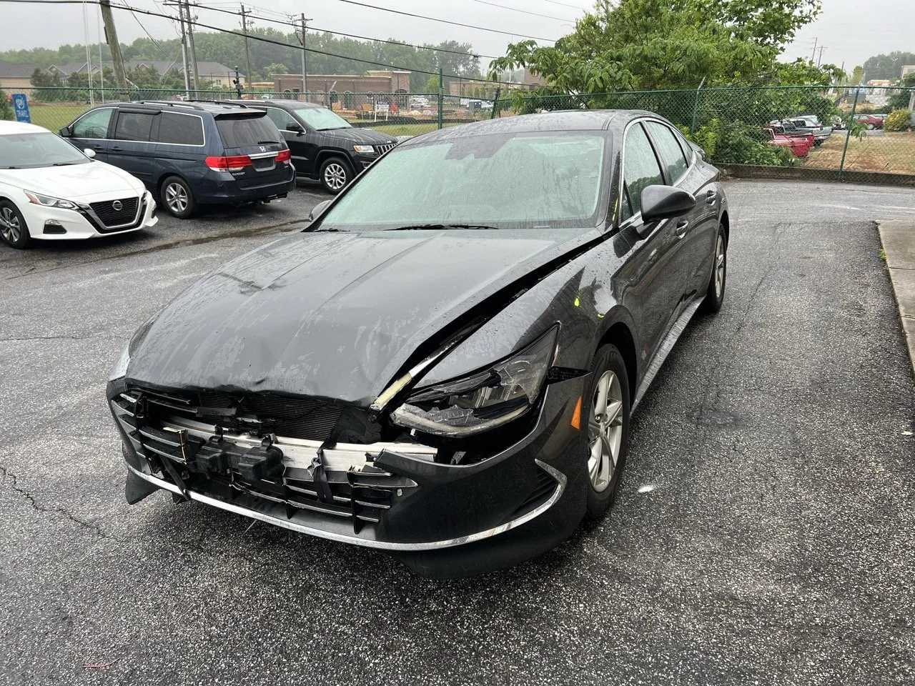 Black sedan car with front-end damage, missing grille, hood crumpled, and parts hanging, parked on wet asphalt in a parking lot.