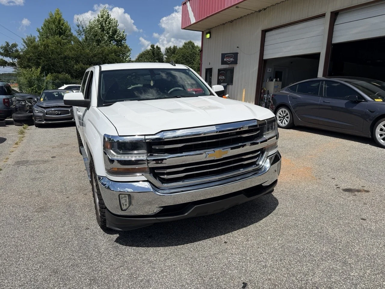 Front view of a white Chevrolet pickup truck parked outside a car dealership, with other vehicles and a building in the background.