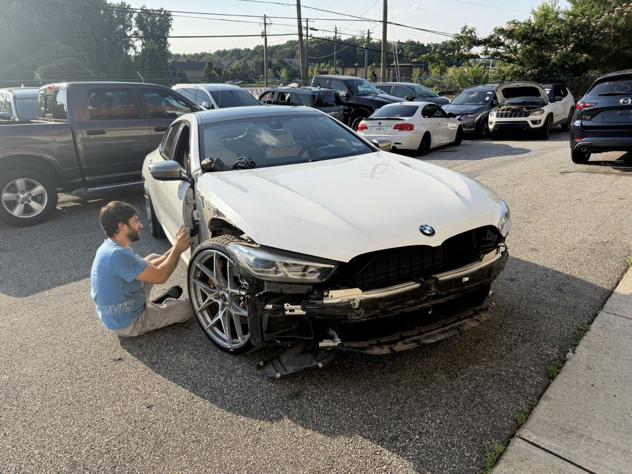 A man sitting on the ground near a silver BMW with front damage, working on the car in a parking lot filled with other vehicles.