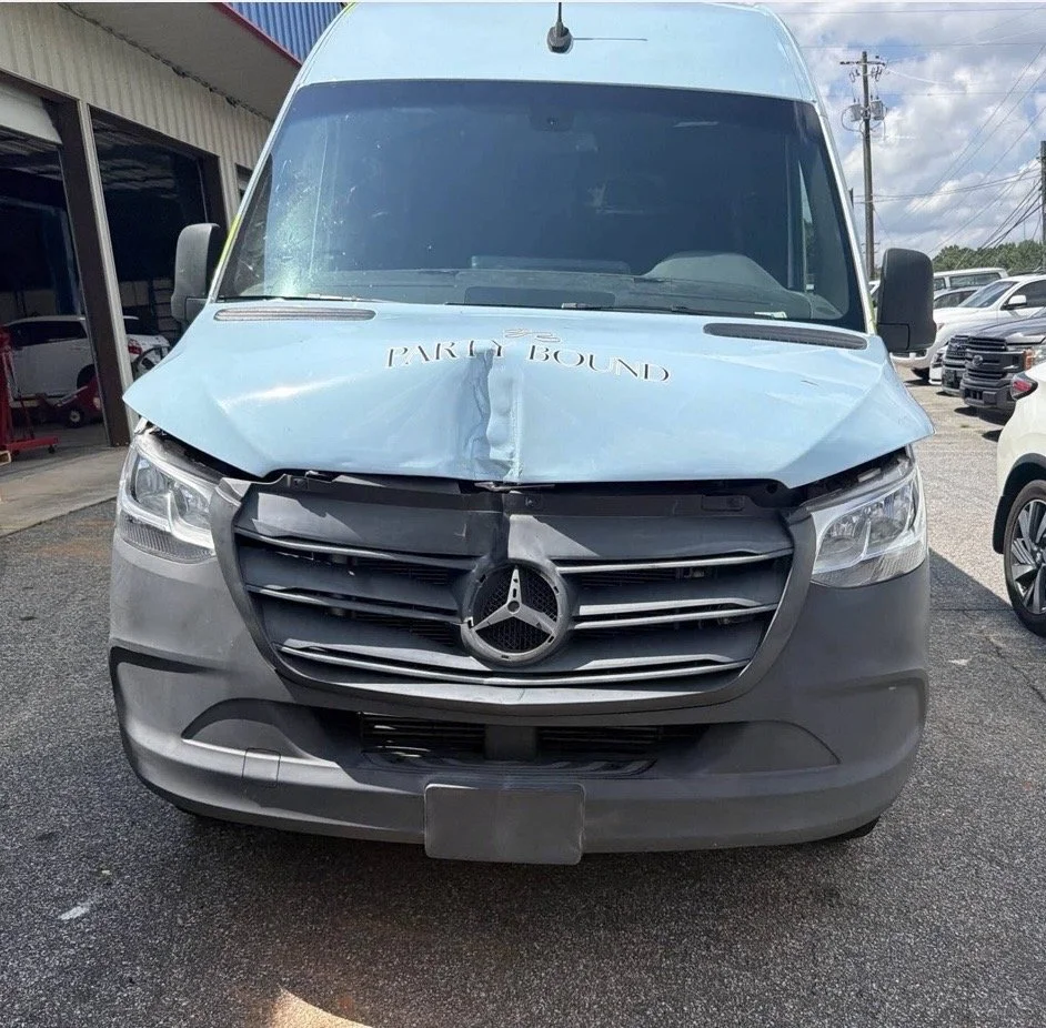 Front view of a damaged Mercedes-Benz van with a crumpled hood and a drawn-on sign that says 'PARTS BOUND' on the windshield, parked in a lot with other vehicles in the background.