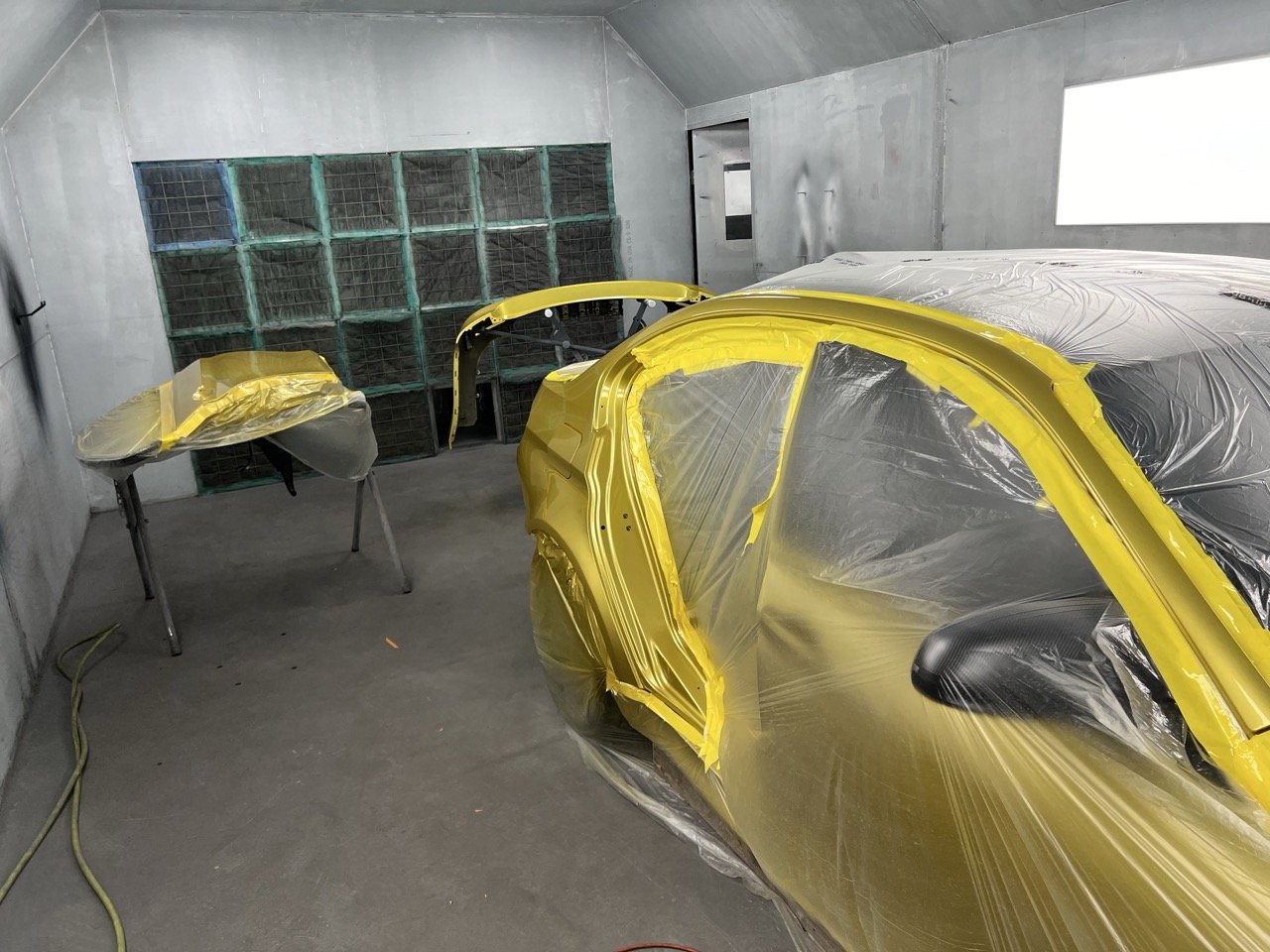 A car being painted in a workshop, covered with plastic sheeting and yellow tape, with parts on stands and a wall of storage boxes in the background.
