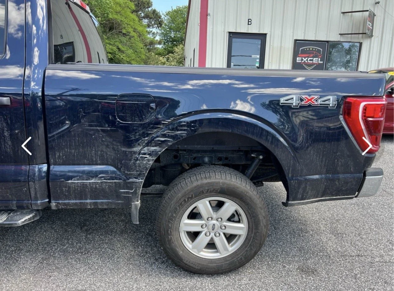 A dark blue pickup truck with visible scratches and dirt on the side, parked outdoors on asphalt. The rear tire is visible, and there is a logo '4X4' on the bed. In the background, there is a white building with a door, window, and signs, including one that says 'Excel.'
