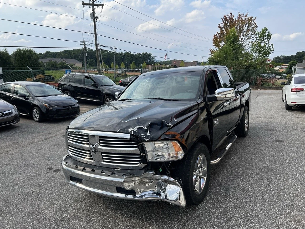 Black Dodge Ram pickup truck with front-end damage, parked in a car lot with other vehicles and trees in the background.