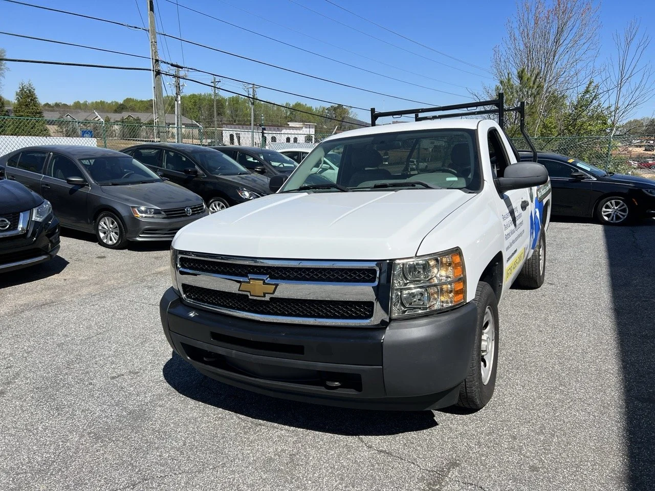 White Chevrolet utility truck parked in a lot with other cars, black bumper, and a ladder rack on top.
