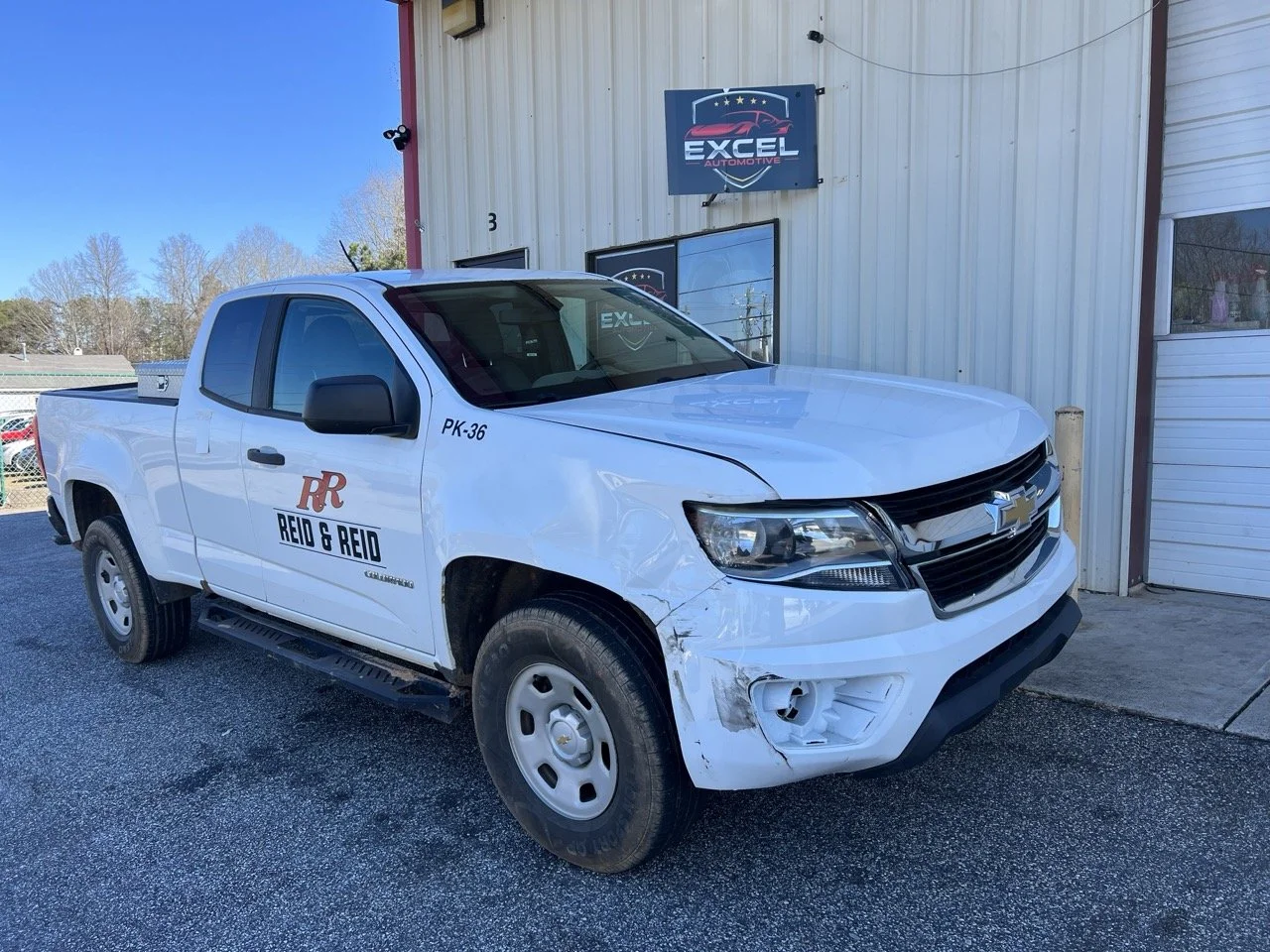 White Chevrolet pickup truck with damage on the front right side, parked outside a building with a sign that reads "Excel Automotive." The truck has "Reid & Reid" and the initials "RR" on the door.