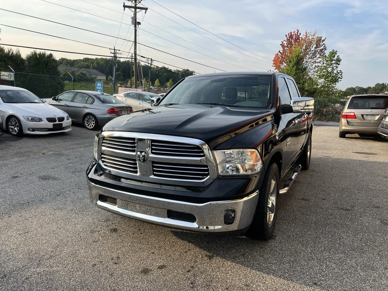 Black Dodge Ram pickup truck parked in a lot with other vehicles, chain-link fence, power lines, and trees in the background, during daylight.