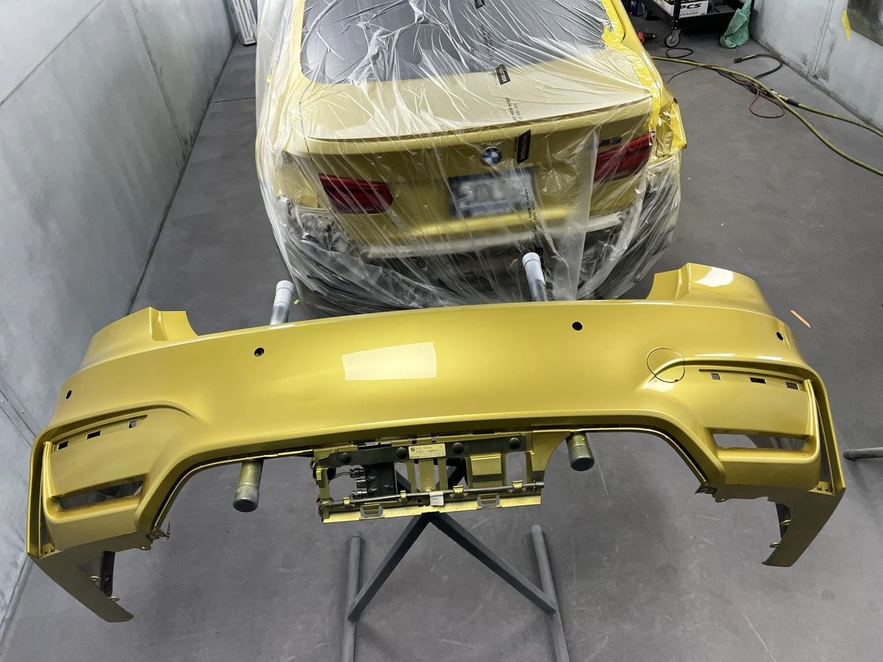 A yellow BMW car front bumper and rear car body are in a garage or workshop. The rear car is covered with protective plastic and is partially covered with a yellow film, suggesting they are being painted or assembled.