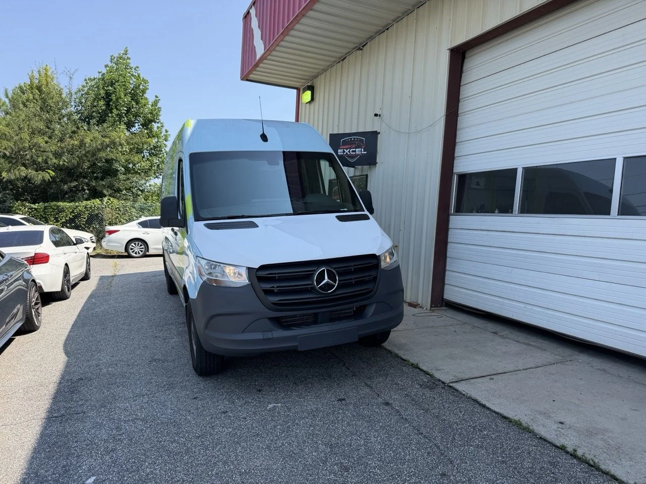 A white Mercedes-Benz van parked outside a building with a white garage door and a sign that reads 'Excel.' There are several other cars parked nearby, and trees are visible in the background.