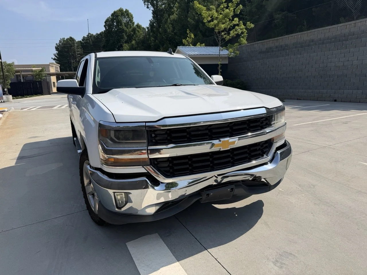 Front view of a white Chevrolet Silverado pickup truck with damage to the front bumper, parked on a concrete lot with trees and a brick wall in the background.