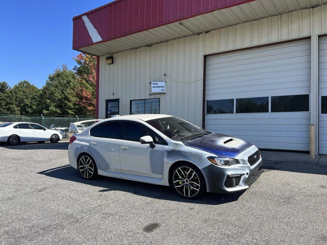A silver and dark blue Subaru WRX with tinted windows parked outside an automotive shop called Excel Automotive LLC, with other cars and a green fence in the background under a clear blue sky.