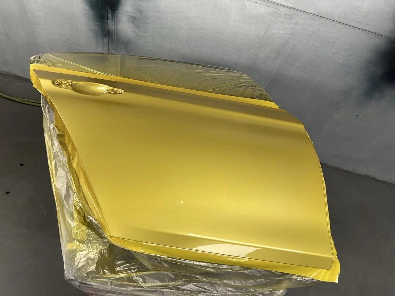 A car hood painted bright yellow, placed in a spray booth with protective plastic wrapping underneath.