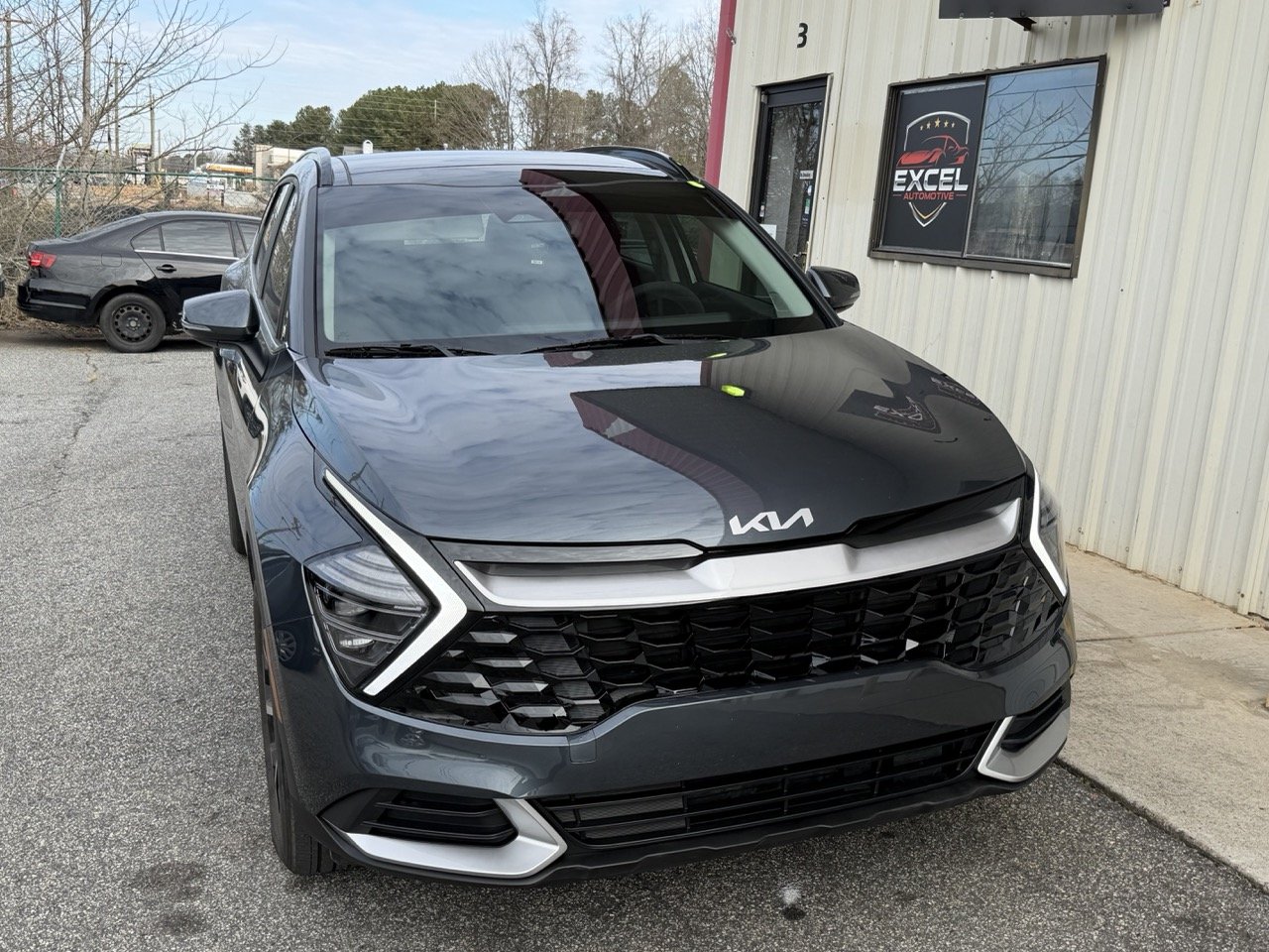 A black Kia SUV parked outside a building with a sign that reads Excel Automotive, with another black car parked in the background.