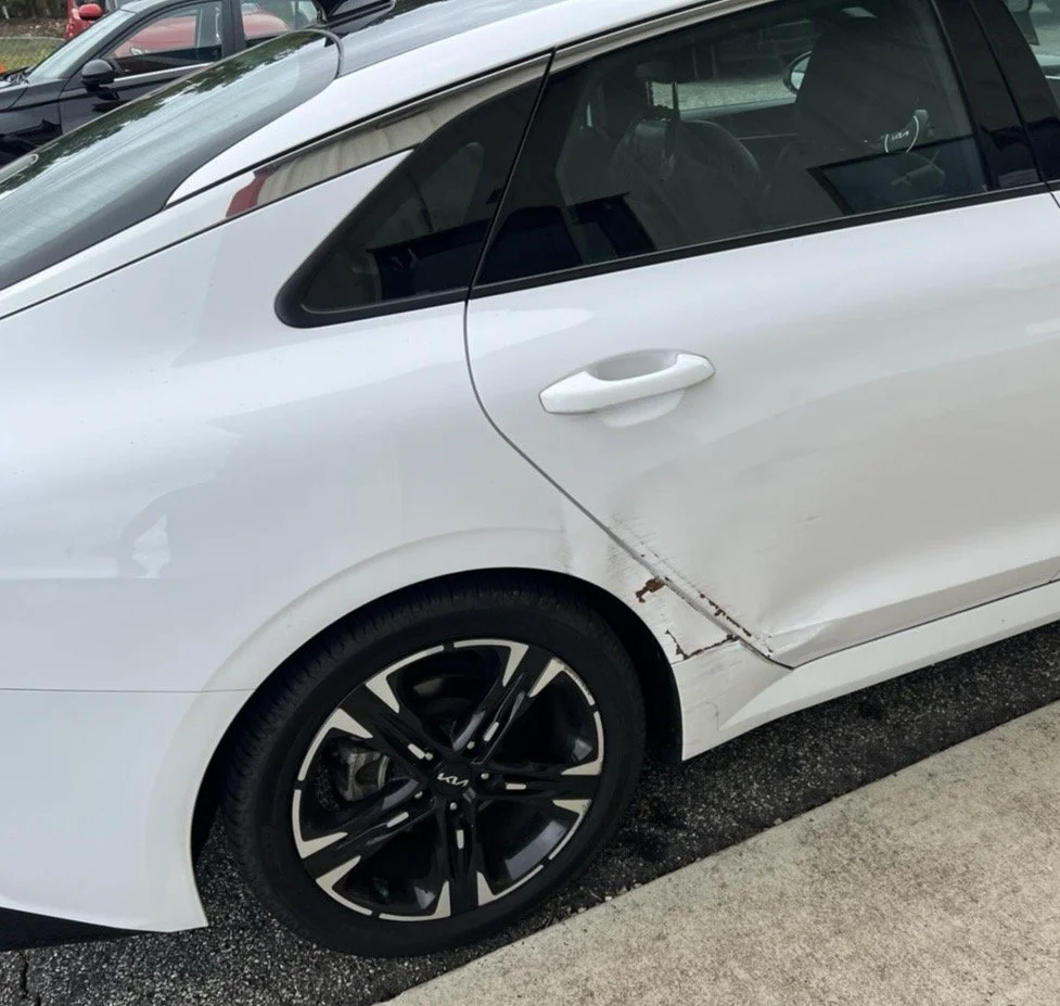 White car with visible damage and rust on the lower side of the rear door and panel, parked in a lot.