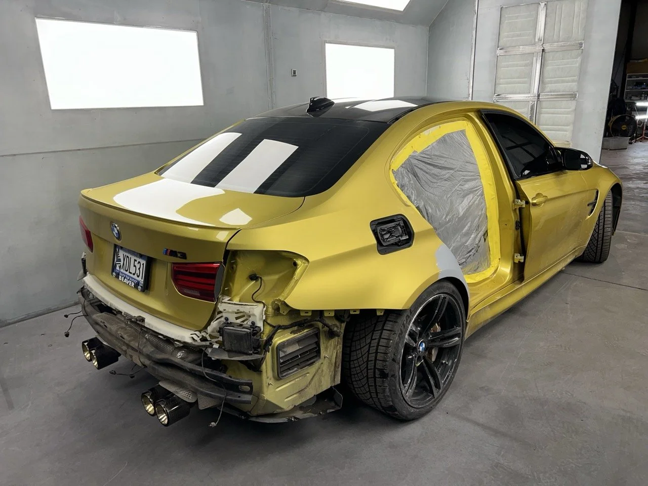 A yellow BMW M3 car in a garage undergoing repairs or customization, with the rear bumper and wheel well area partially disassembled and the rear light and wheel intact.