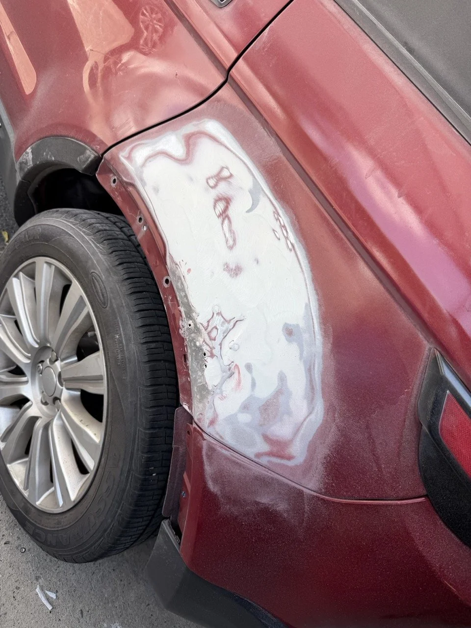 Red car with a missing front fender, exposing the wheel and inner workings; the area around the wheel is rough and unpainted.