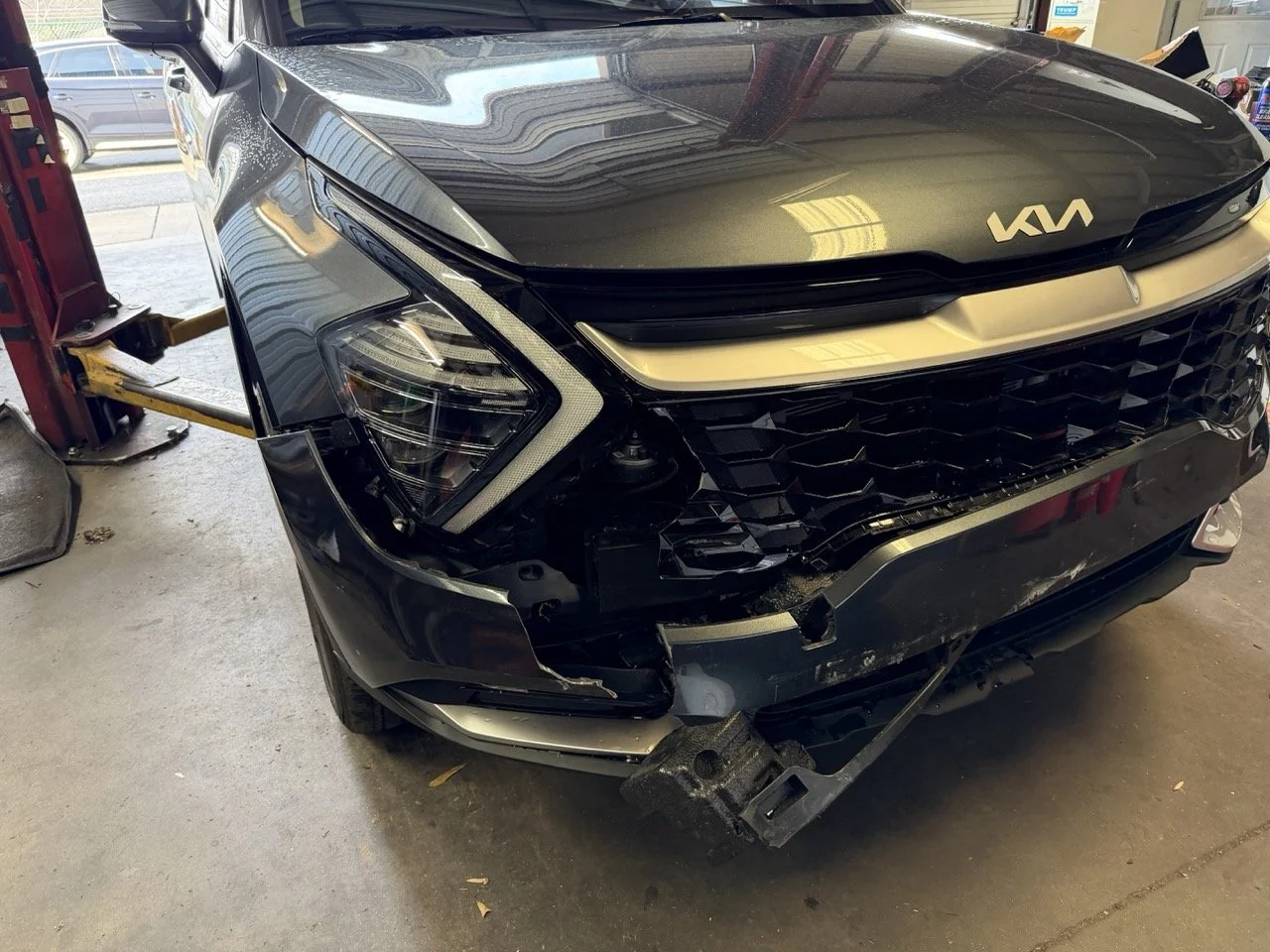 Front view of a black Kia vehicle with significant front-end damage, appearing to be in a repair shop.